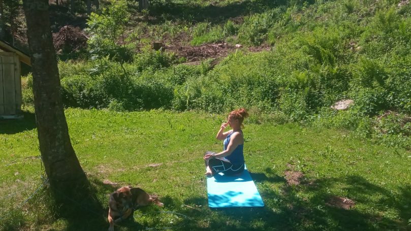 Frau sitzt im Freien auf einer Matte und meditiert, umgeben von Grün und Bäumen.
